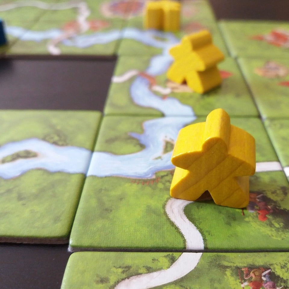 Wooden board game pieces on cardboard tiles depicting fields, rivers, and towns